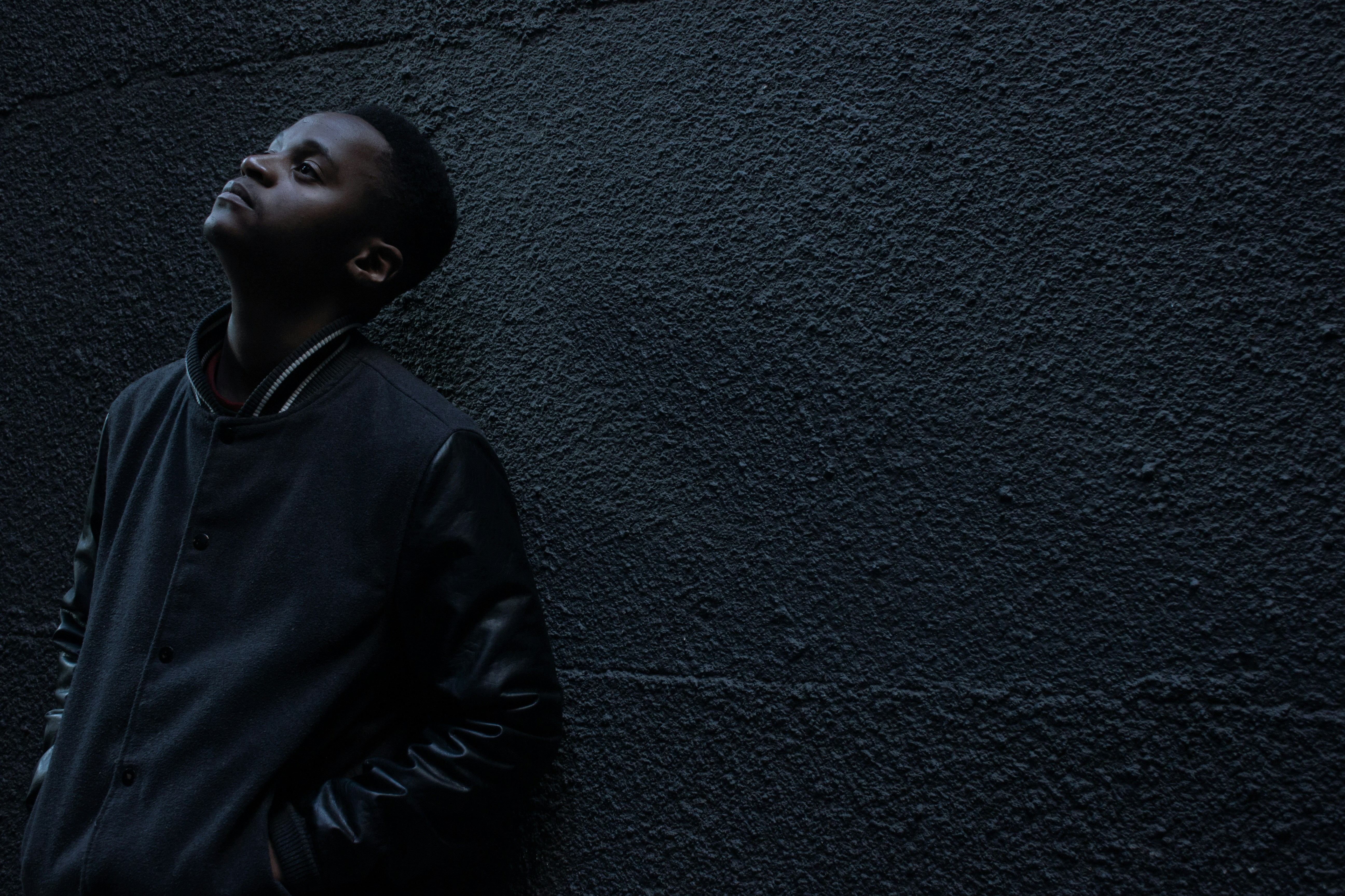 man wearing black button-up jacket standing and leaning against the wall while looking up